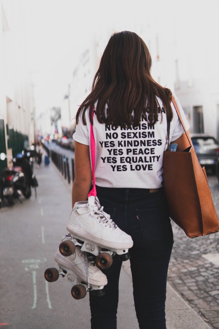 a lady with skating shoes hanging on her shoulders while wearing a t-shirt with the caption; no homophobia, no racism, no sexism, yes kindness, yes peace, yes equality, yes love